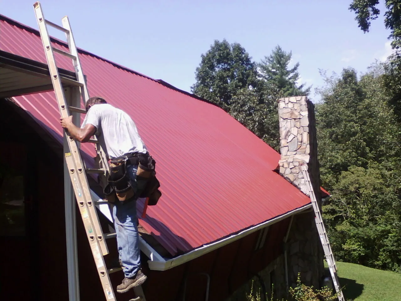 Roofer on a ladder beside a red metal roof and stone chimney — Soffit & Fascia Repair by Roofing Brothers in Stahlstown